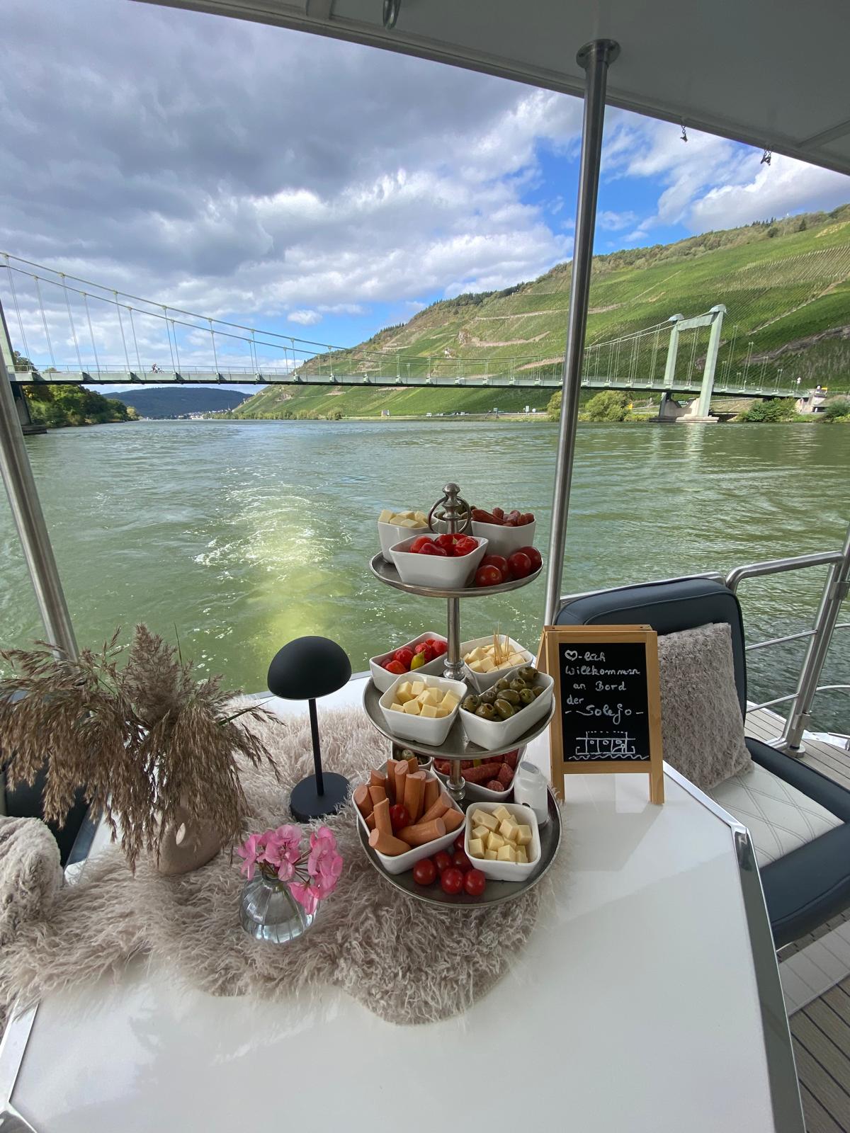 Willkommenstafel mit Fingerfood und Willkommensschild an Bord der SOLEJO, dahinter Mosel und Hängebrücke